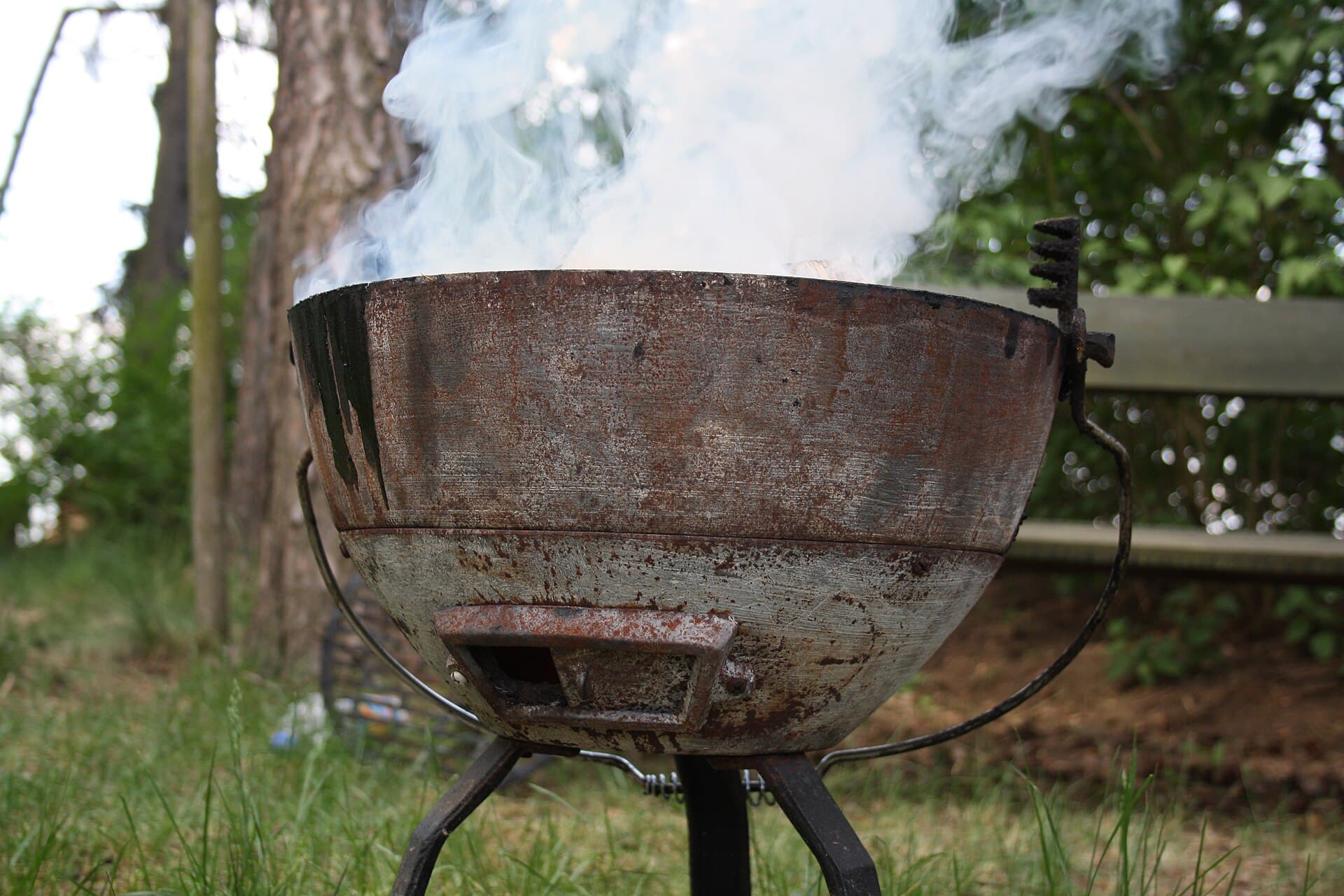 A weathered kettle charcoal barbecue sitting on grass with a plume of white smoke rising from the bowl