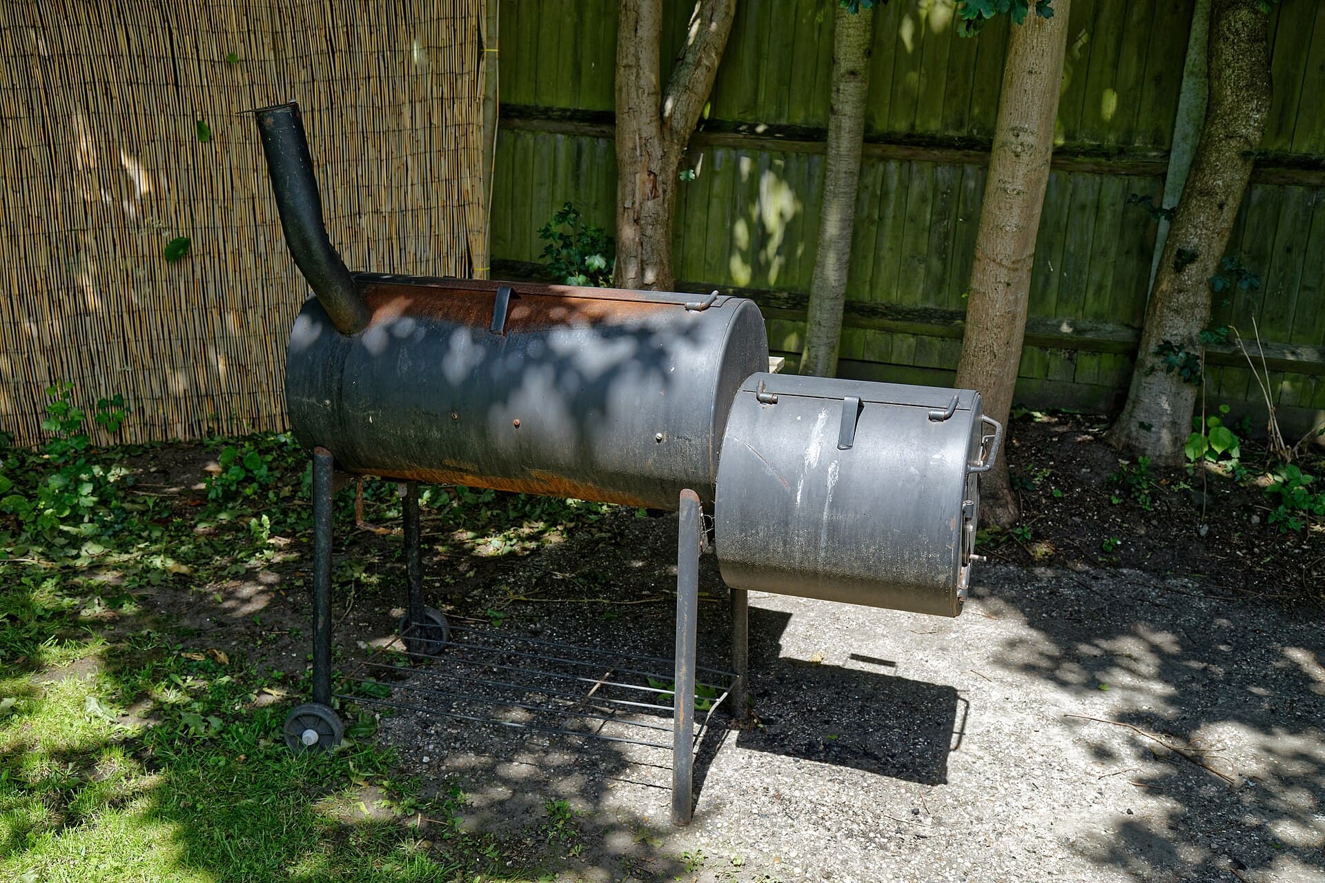 A black offset smoker on a wheeled stand in a backyard, with a timber fence and tree trunks behind it