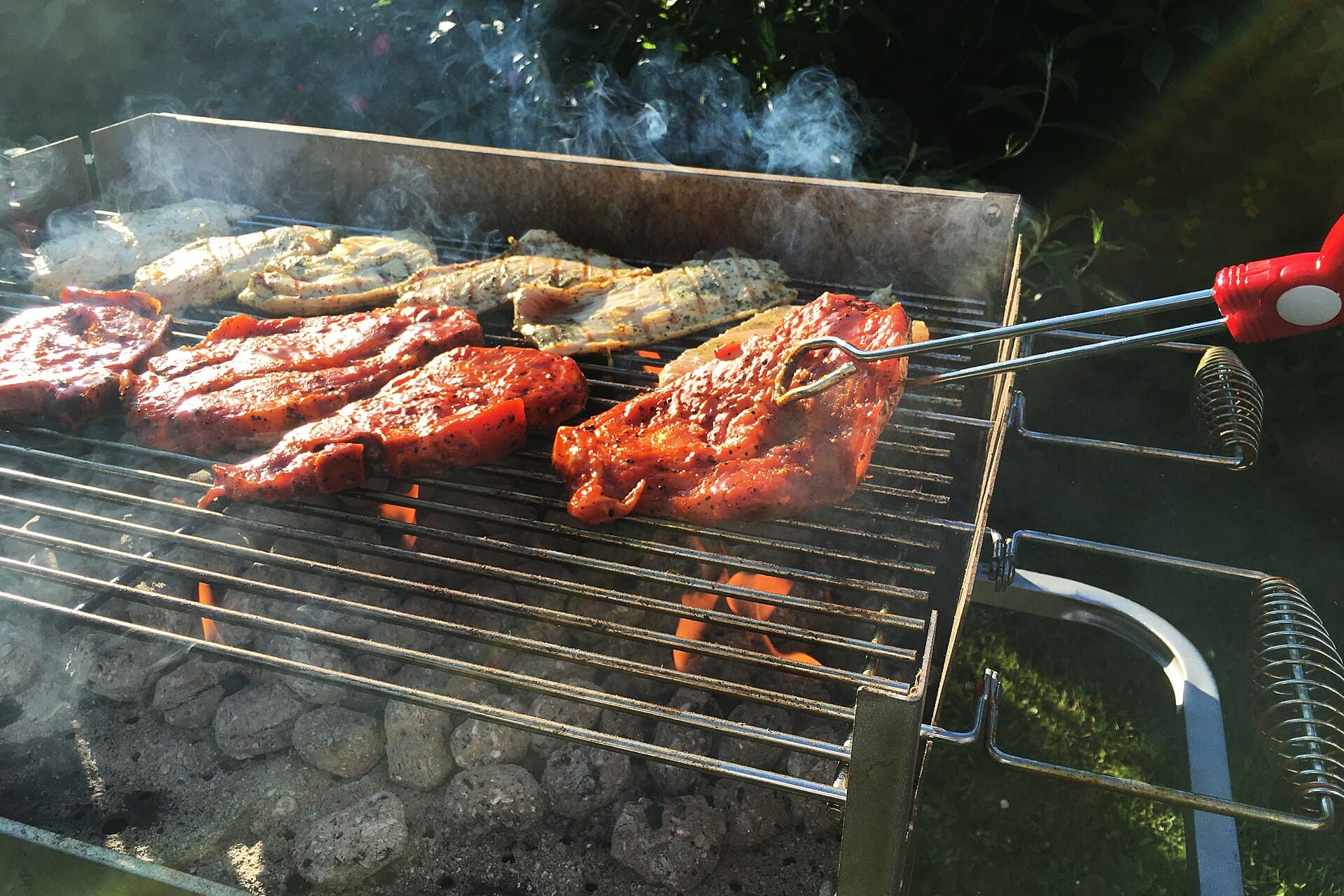 Marinated steaks and pale fillets cooking over glowing charcoal, with red-handled tongs reaching toward the meat
