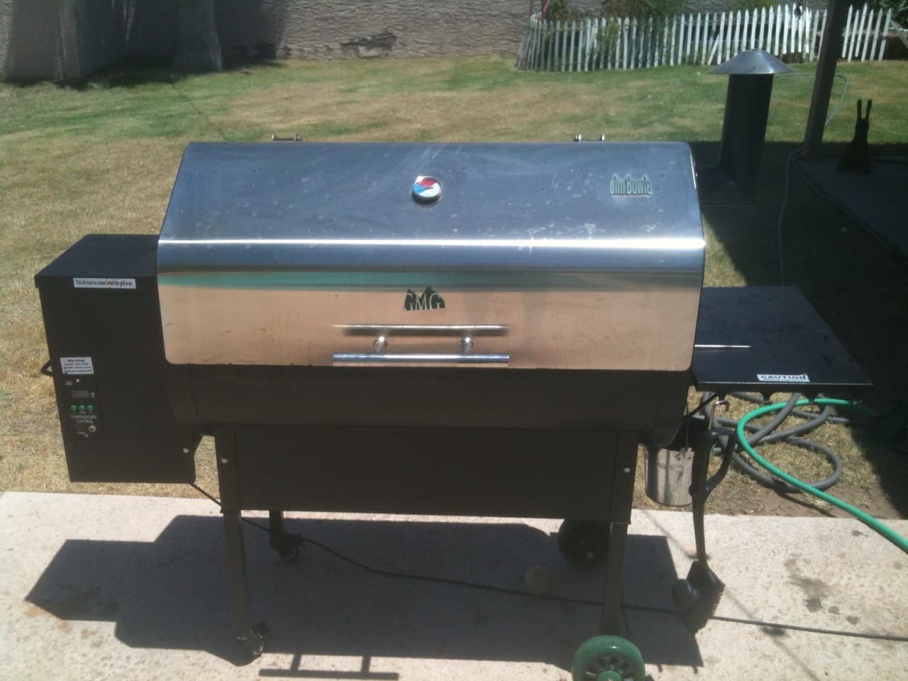 A stainless-lid pellet barbecue on a black trolley with a hopper on the left and a small side shelf on the right, standing on a concrete pad in a backyard
