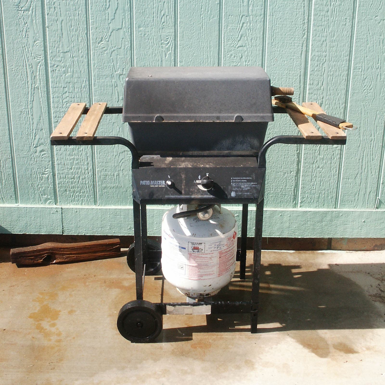 A black hooded gas barbecue on a wheeled trolley with a white LPG cylinder, wooden side shelves and a mint-green weatherboard wall behind it