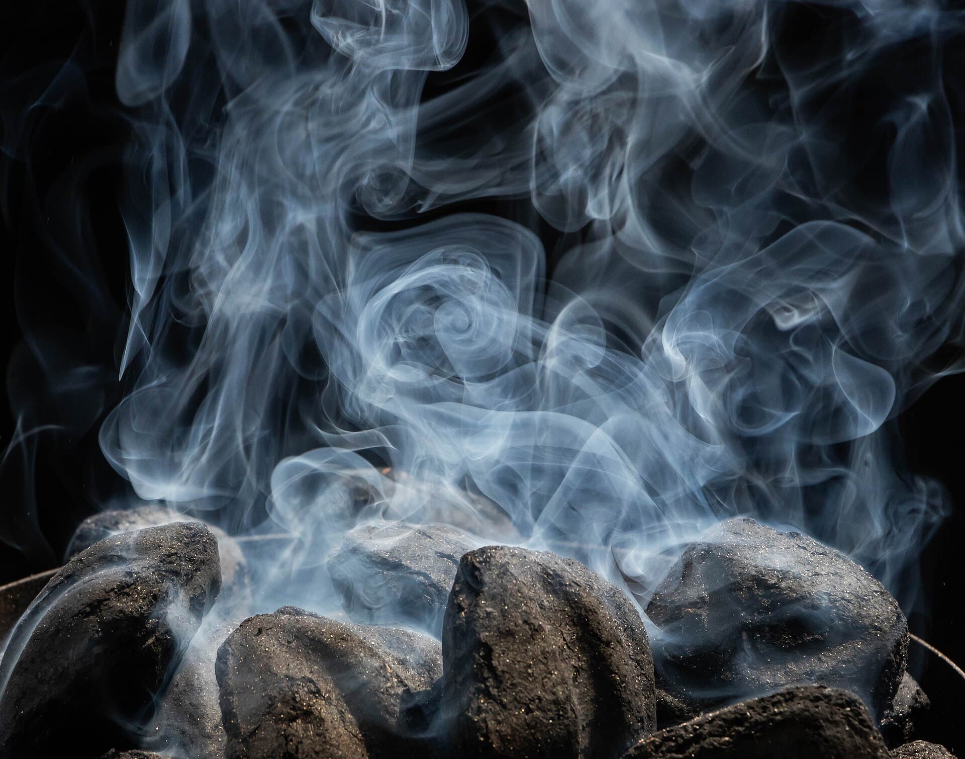 Close-up of charcoal briquettes with pale smoke curling upward against a black background