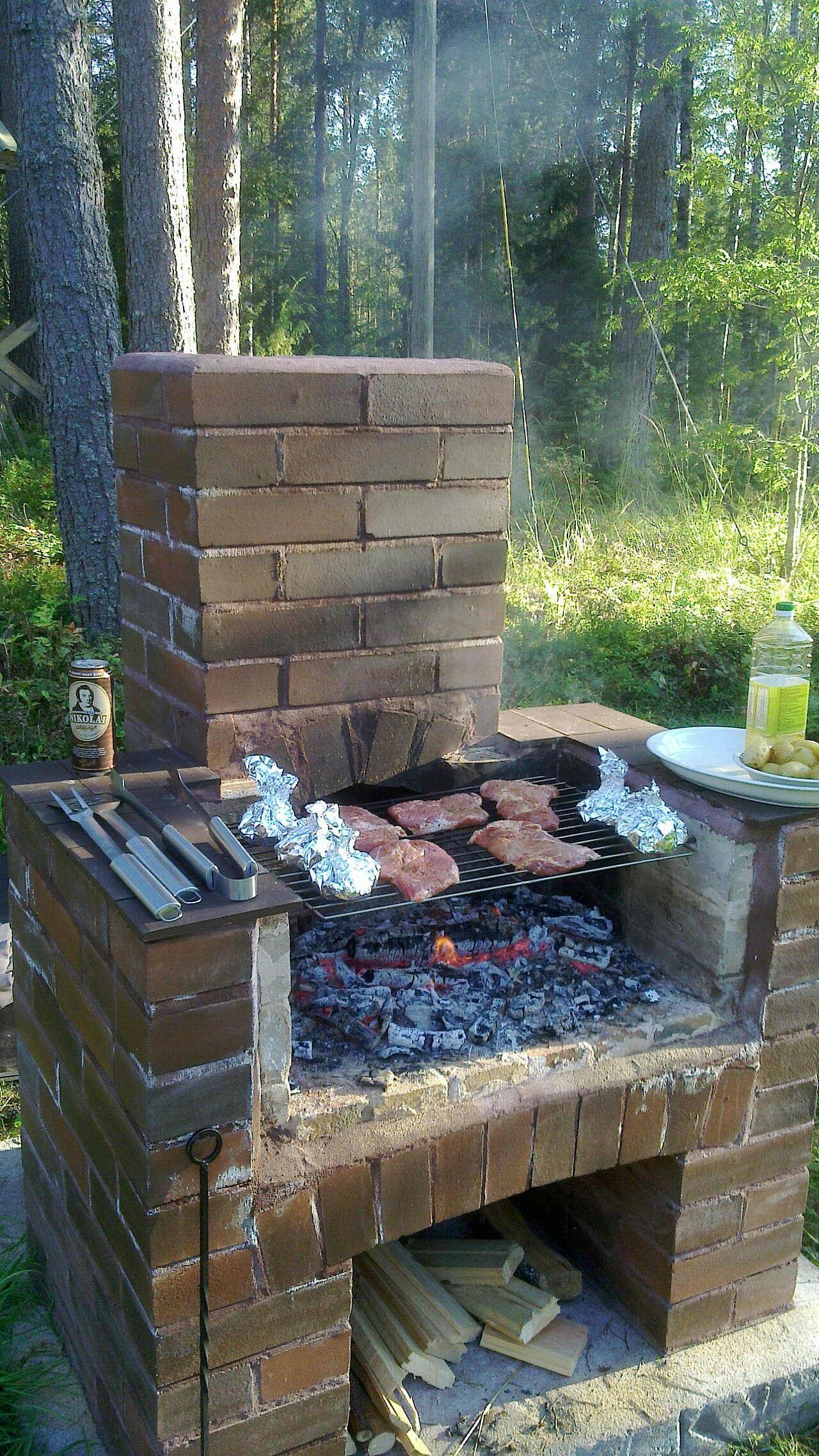 A red-brick outdoor barbecue with a chimney, meat and foil parcels on the grate over glowing coals, and split firewood stacked in the alcove below